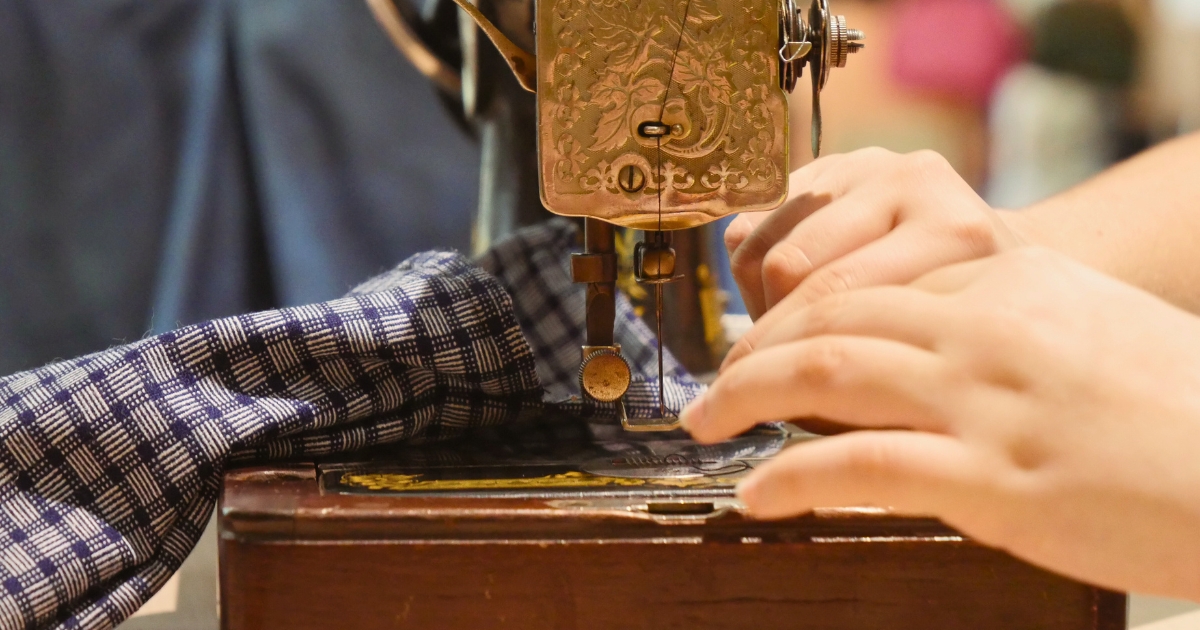 Gros plan sur une machine à coudre ancienne en fonctionnement. Des mains cousent un tissu à motifs bleus, mettant en valeur un savoir-faire artisanal et le travail du textile.